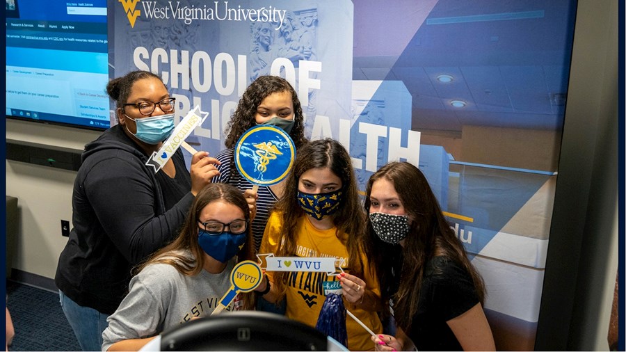 Five new School of Public Health students pose for a group photo as they stand in front of a School of Public Health vertical banner, holding WVU photo props that say I love WVU and I love Public Health