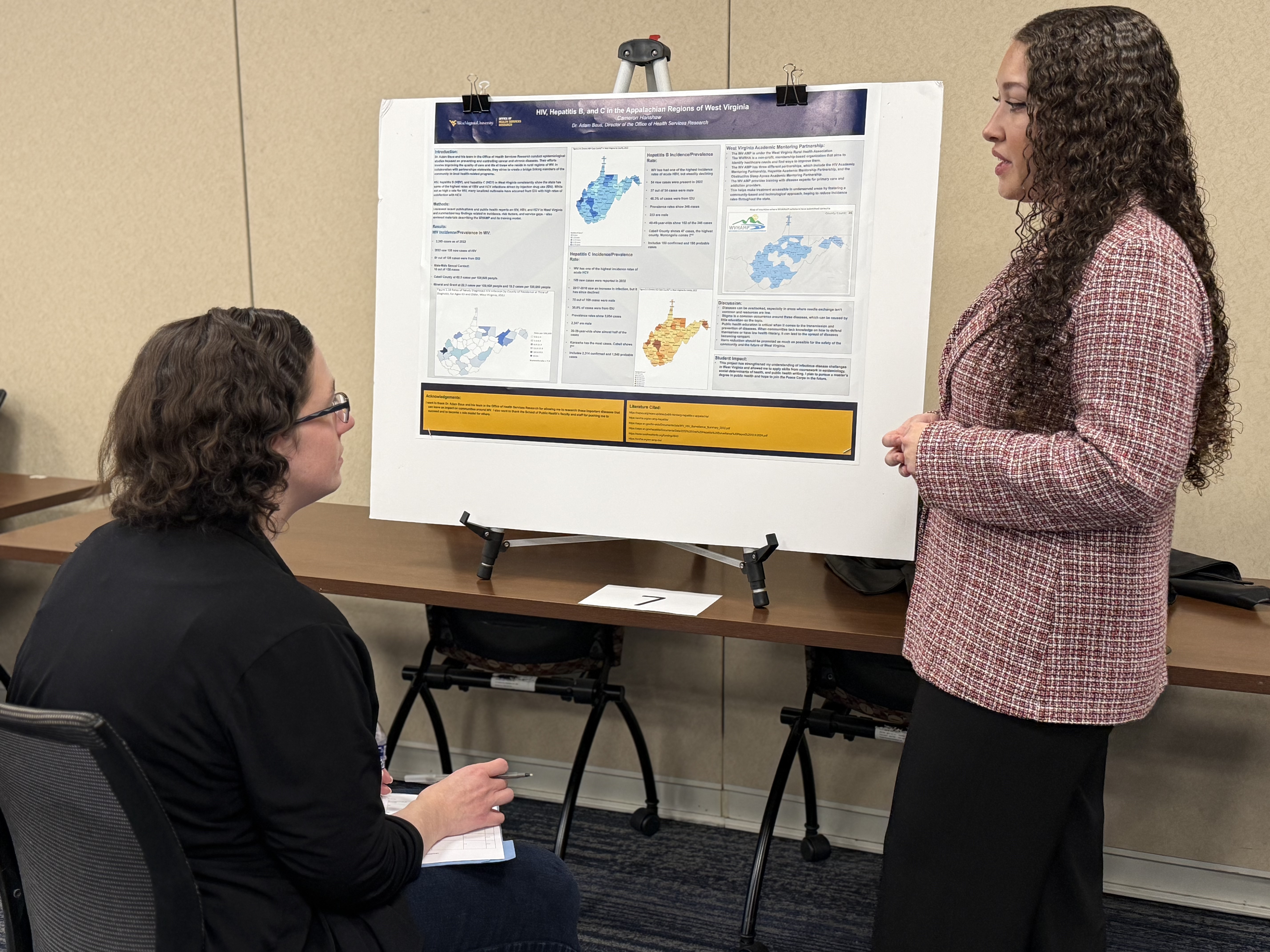 Student stands beside a research poster giving a presentation to individual seated in front of her. 