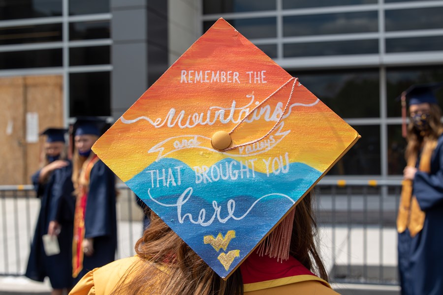 Public Health graduate stands with back to camera showing off decorated graduation cap, which reads "Remember the mountains and valleys that brought you here"