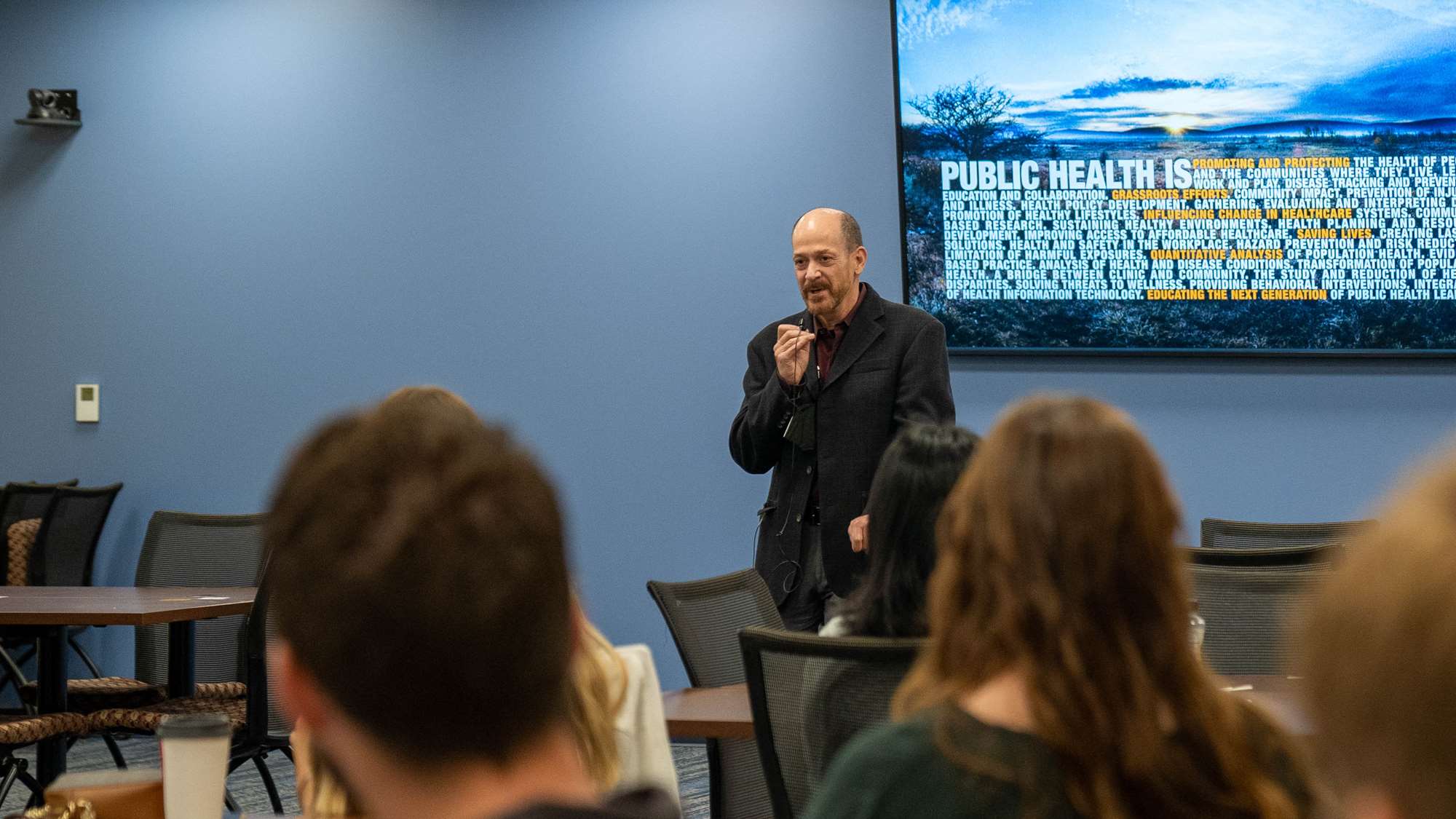 Dean Coben stands in a room of students holding a lavalier mic, with a large wall-mounted monitor behind him that reads Public Health is, followed by a variety of short phrases describing public health. 