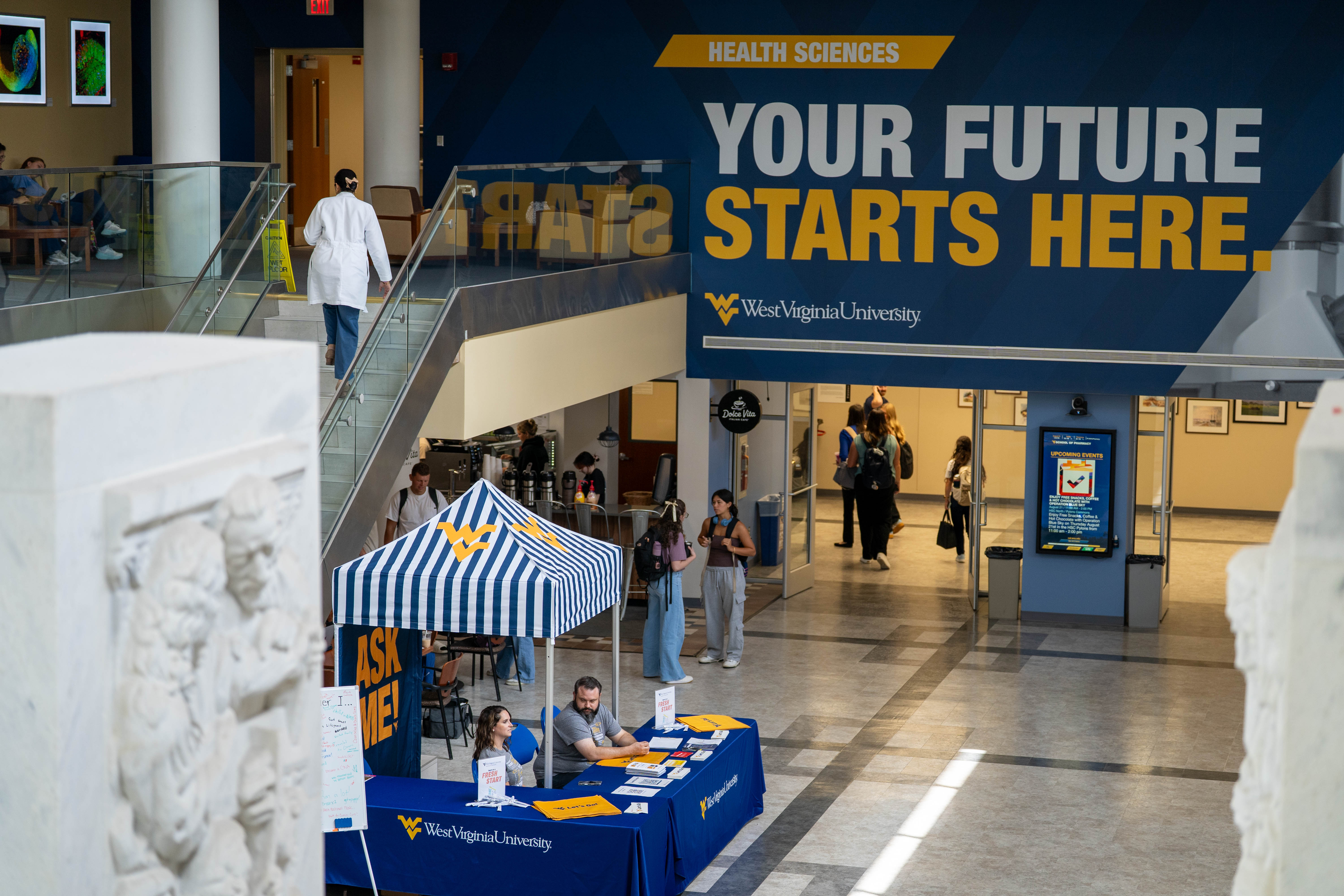 Wall mural above doorway of over large open foyer. Mural has photo of person standing on mountain holding Let's Go flag with sunset in background. Mural reads: WVU Health Sciences. Your future starts here. Let's Go! 