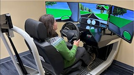 Dr Rudisill is how seated in a chair with multiple monitors in front displaying driving imagery.